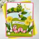 A handmade children's greeting card featuring layered paper crafts of two cute cartoon caterpillars, one green and one pink, along with yellow flowers, grass, and a banner that reads 'JUST SAVING HELLO'. The card is displayed on top of yellow and green envelopes against a light gray background with soft white flowers in the corner.