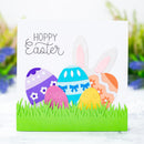 A square Easter greeting card with a white background, featuring the words “HOPPY Easter” above four colorful patterned eggs in a green grass die-cut design. The card is photographed against a softly blurred outdoor background with greenery and flowers.