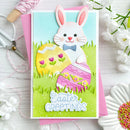 A handmade Easter greeting card featuring a white bunny with pink ears, a blue bow tie, and two patterned Easter eggs on a green grassy background. The card is layered over a pink envelope and styled on a white wooden surface with scissors, thread, flowers, and sprinkles.