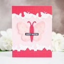 A handmade Easter greeting card featuring a pink and red butterfly design, white lace-like borders, and a black banner reading 'HAPPY EASTER', photographed against a softly blurred background with white flowers.
