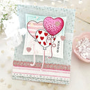 A handmade layered greeting card featuring two heart-shaped balloons, one pink glitter heart and one white heart with red hearts, on a light blue patterned card base. The card is styled on a beige fabric background with decorative props.
