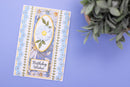 A handmade greeting card with a floral daisy design and the words 'Birthday Wishes' on a lavender background, styled next to a green succulent plant.