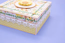 A decorative gift box with a floral patterned lid, white lace trim, and a yellow flower topper, set against a solid light purple background.