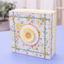 A decorative square gift box with a white background and a colorful floral print, featuring a large yellow flower applique on the front, set on a light blue surface.