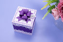 A square gift box in purple and white with a large purple bow on top, decorated with a 'Happy Birthday' tag. The box is set on a light blue surface next to a vase of pink roses.