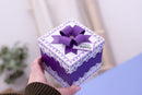 A hand holding a square purple and white gift box decorated with a matching purple bow, lace trim, and a small 'Happy Birthday' tag, against a softly blurred indoor background.