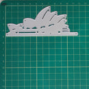 A silver metal die-cut template shaped like the Sydney Opera House, displayed on a green cutting mat with grid lines.