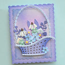 A handmade layered card featuring a lavender and purple basket filled with 3D die-cut flowers in shades of blue, green, white, and yellow, set against a light mint green background.