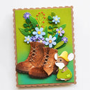 A handmade paper craft card featuring a pair of brown lace-up boots, a small mouse in green clothing and a yellow hat, and blue and purple flowers with green leaves on a green background.