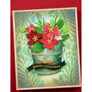 A handmade Christmas greeting card featuring red poinsettia flowers and green leaves arranged in a distressed teal metal bucket, with a black banner reading 'MERRY CHRISTMAS' on a patterned cream and green background, set against a dark red backdrop.