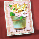 A handmade layered greeting card featuring a scalloped pink and peach border, a green and gold watering can, and layered paper flowers and leaves with the words 'thank you' in gold foil lettering. The card is photographed flat on a textured dark red background.