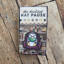 A rectangular decorative card or bookmark with German text reading 'Die Realität HAT PAUSE' at the top, featuring an illustration of a penguin sitting in a chair and reading a green book. The item is photographed on a rustic wooden surface.