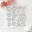 A square clear plastic or silicone stamping/cutting template sheet with floral and leaf outlines, photographed on a white surface with pink flowers in the top left and a small white decorative object in the bottom right.