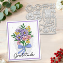 A handmade greeting card with a floral bouquet illustration and the word 'Gratitude' is shown on a light wood surface alongside metal die-cutting templates. The card has a lavender border and white center, with purple, pink, yellow, and green flowers and a blue ribbon.