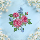 An embroidered floral design featuring two pink roses with green leaves and small blue flowers, arranged on a light blue marble background with white baby's breath flowers framing the edges.