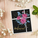 A square greeting card with a black background, pink and blue flowers, and the word 'believe' in gold script, styled on a light wood surface with white baby's breath and mint leaves.