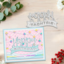 A handmade layered card with a pastel pink and blue design, shown alongside a matching metal die-cut stamp on a light wood background with decorative eucalyptus and red berries.