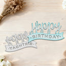 A decorative metal 'Happy Birthday' sign shown in two color options on a light wood background with dried flowers and small white blossoms. One sign is silver and the other is light blue, both with ribbon banners and cursive script lettering.