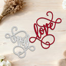 Two decorative metal word cutouts, one red and one silver, on a wooden surface with flowers.