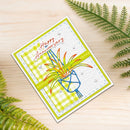A handmade square greeting card with a 'Happy Anniversary' design, styled on a light wood surface with green fern leaves. The card features a white textured background, a lime green and white diagonal plaid border, a bright lime green horizontal stripe, and a blue geometric flower holding orange and yellow petals.