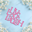 Pink laser-cut wooden sign spelling out 'Live Laugh Love' with a heart symbol, displayed on a light blue marble background with white baby's breath flowers.