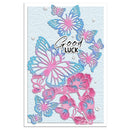 A square greeting card featuring layered die-cut butterflies and flowers in pink and blue on a light blue textured background, with the words 'Good Luck' in white script and small clear rhinestone accents.