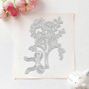 A floral die-cut stamp or embosser shown on a square sheet of matching light pink paper, with pink roses in the top-left corner and a small white ceramic object in the bottom-right on a white background.