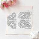 A butterfly-shaped metal craft die set displayed on a square sheet of white cardstock, styled with pink roses in the top-left corner and a small white ceramic figurine in the bottom-right on a white background.