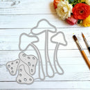A metal die-cut craft file shaped like mushrooms, displayed on a white wooden background with paintbrushes and flowers in the corner.