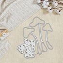 A mushroom-shaped metal cutting die set with a silver-colored outline, displayed on a textured cream fabric background alongside a waffle-knit textile, cotton stems, and a small wooden-and-white camera accessory.