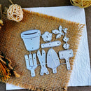 A set of metal garden-themed die-cut shapes, including a watering can, flower, leaves, and gardening tools, arranged on a burlap fabric over a white textured paper background.