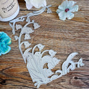 A decorative metal die-cut sheet with floral and bird motifs, displayed on a rustic wooden surface alongside flowers and a candle jar.