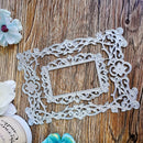 A silver metal die-cut decorative frame with intricate scrollwork and floral motifs, displayed on a rustic wooden surface with decorative flowers.