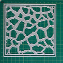 A square metal stencil with an organic, web-like cutout pattern, displayed flat on a green cutting mat with grid lines.