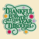 A decorative typographic sign reading “THANKFUL FOR YOU ALL THE YEAR THROUGH” in layered green lettering with a pale mint outline, accented by small yellow and pink flowers on a light cream background.