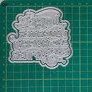 A silver metal die-cut craft embellishment with the words 'LOVE LAUGH AND HAPPY EVER AFTER' in decorative lettering, shown on a green cutting mat with grid lines.