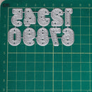 A set of metal letter cutters in the shape of stylized numbers and letters, arranged on a green cutting mat with grid lines.
