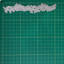 A silver metal die-cut floral border with leaf and vine patterns, displayed on a green cutting mat with centimeter grid lines.