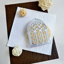 A silver metal die-cut window frame ornament lies on a textured white paper square, placed on a dark brown wood board with decorative cream yarn balls around it.