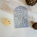 A silver metal die-cut ornament with an intricate filigree pattern, displayed on a light beige textured surface with decorative woven balls.