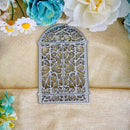 An ornate silver metal arch-shaped frame or decorative cutout with intricate scrollwork, displayed on a beige fabric background with artificial flowers around the edges.