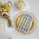 A silver metal decorative arch-shaped ornament with an intricate lattice pattern, displayed on a round woven straw plate. The setup is styled as a flat-lay with dried flowers, a yellow pom-pom, and a white textured sheet on a neutral background.