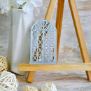 A silver metal window frame die-cut stamp or ornament displayed on a wooden easel stand, with decorative white flowers in a vase and woven balls in the background.
