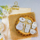 A set of silver metal enamel pins shaped like coffee and bakery items, displayed on a round woven tray over a burlap cloth with decorative flowers and a gold binder clip.