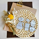 A set of coffee-themed paper cutouts, including a coffee pot, takeaway coffee cup, cupcake, and mug, arranged on a round woven placemat with dried flowers and a brown paper clip on a light background.