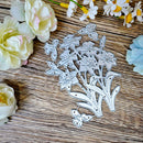 A floral metal cutting die with intricate flower and leaf shapes, displayed on a rustic wooden surface surrounded by decorative white and pale yellow flowers.