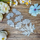 A set of silver metal floral die-cut craft stamps or cutters arranged on a rustic wooden surface, surrounded by decorative paper flowers in white, pale pink, light blue, and purple.