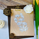 A botanical floral stamp or stencil design shown on a brown kraft paper sheet clipped to a dark board with a gold binder clip, styled with decorative twine balls, a white flower, and green leaves on a wood and light blue background.