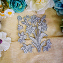 A floral metal die-cut craft template featuring daffodils and buttercups, displayed on a textured beige fabric background with decorative paper flowers.