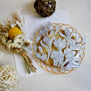 A floral die-cut sticker or decal sheet with silver-white botanical outlines displayed on a round gold wire tray, styled with dried flowers, a yellow pom-pom, and woven decorative balls on a light beige textured background.